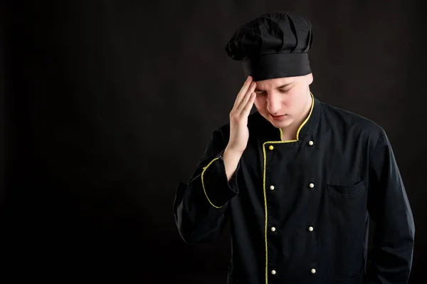 Portrait of young male dressed in a black chef suit with open arms looking up, has headache posing on a black isolated background with copy space advertising area