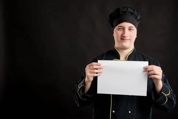 Portrait of young male dressed in a black chef suit holding a white paper and smileing posing on a black isolated background with copy space advertising area