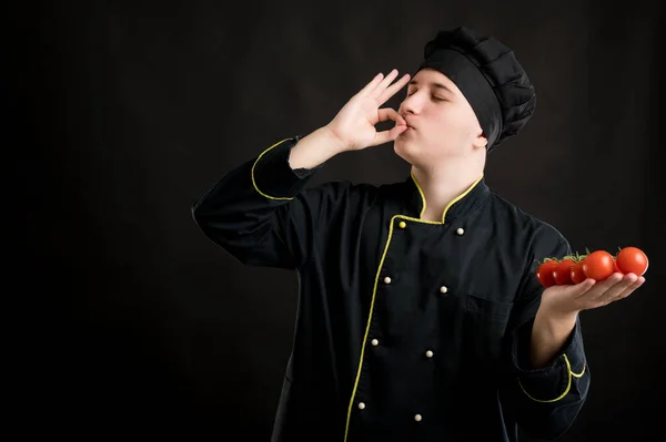 Portrait of young male dressed in a black chef suit holding tomato showing ok sign posing using salt on a black isolated background with copy space advertising area