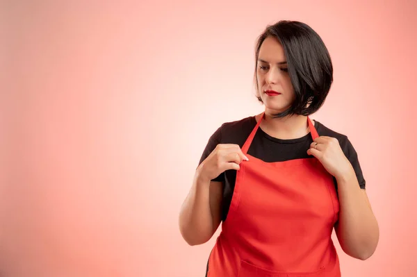 Woman employed at supermarket with red apron and black t-shirt, arrange her apron isolated on red background