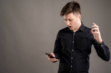 Mobil uygulamalar kullanarak gündelik siyah gömlekli, ciddi ve çekici bir adamın portresi. Gri arka planda izole edilmiş cep telefonuna kızgın yüz ifadesi içeren kısa mesajlar, fotokopi alanı reklam alanı.