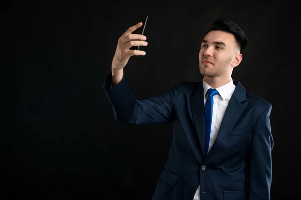 Portrait of angry business man wearing blue business suit and tie doing selfie photo with his cellphone isolated on black background with copy space advertising area