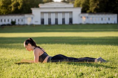 Güzel genç bir kadın sarı bir yatakta yatıyor, parkta dar bir spor kıyafeti giyerken poz veriyor, pilates veya yoga yapıyor, yüzme alıştırmaları yapıyor.