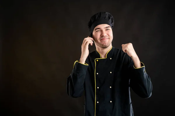 Portrait of young male dressed in a black chef suit with, happy face and gestures and talking on cellphone, posing on a brown background.