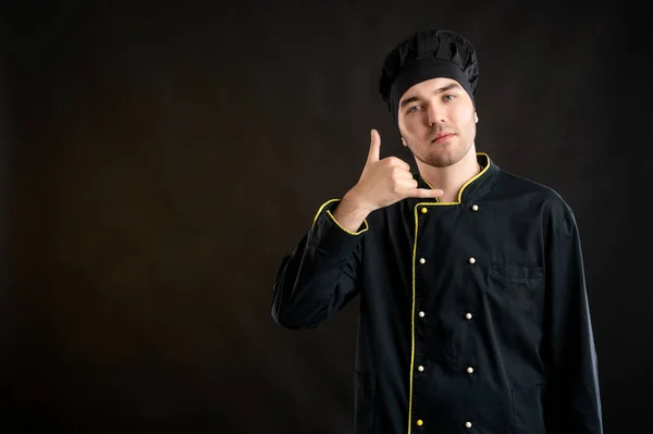 Portrait of young male dressed in a black chef suit showing call me gesture posing on a brown background with copy space advertising area