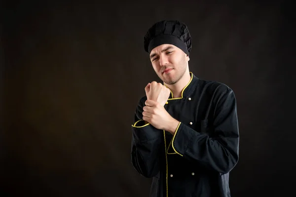 Portrait of young male dressed in a black chef suit has wrist pain posing on a brown background with copy space advertising area