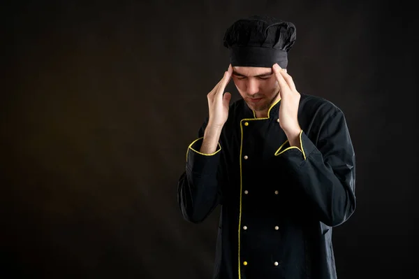 Portrait of young male dressed in a black chef suit with open arms looking up, has headache posing on a brown background with copy space advertising area