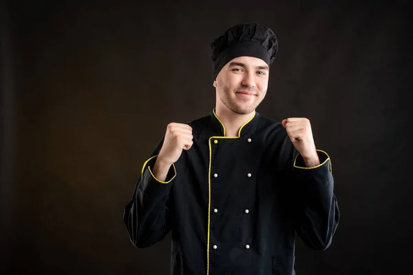 Portrait of young male dressed in a black chef suit raised fists up exclaiming with joy and excitement posing on a brown background with copy space advertising area