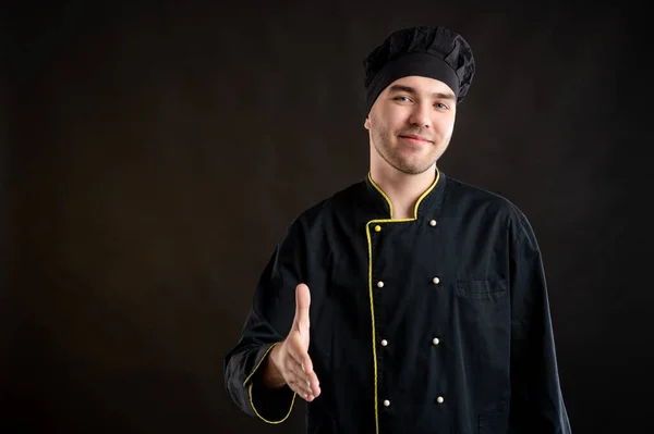 Portrait of young male dressed in a black chef suit shaking hand posing on a brown background with copy space advertising area