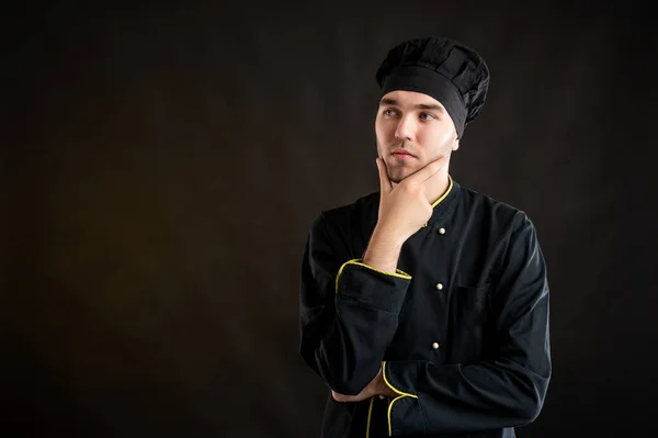 Portrait of young male dressed in a black chef suit holding hand under his chin, having doubtful posing on a brown background with copy space advertising area