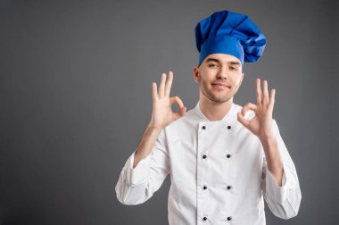 Beyaz şef kıyafeti giymiş genç bir erkek portresi. Gri bir arka planda fotokopi alanı reklam alanı ile çift tamam işareti gösteriyor.