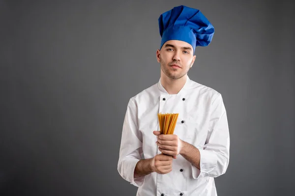 Young male dressed in a white chef suit hold spaghetti in his hand posing on a gray background with copy space advertising area
