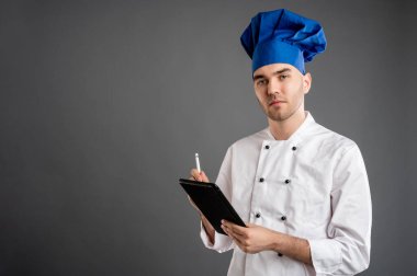 Beyaz şef kıyafeti giymiş genç bir erkeğin portresi notlar alıyor. Gri arka planda fotokopi alanı reklam alanına sahip kameraya bakıyor.