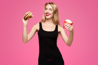 Genç güzel ince kız pembe bir arka plan üzerinde beyaz gülümseme ile hamburger tutar ve patates kızartması onun elinde ve gülümseyerek. Abur cubur kavramı
