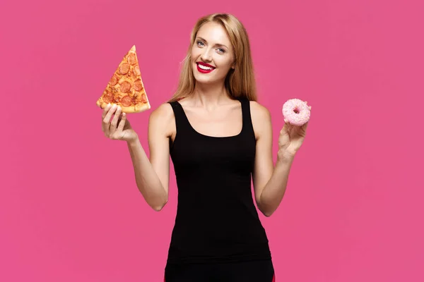 Young beautiful sad girl on a pink background holds pizza and donut in her hands and smiling. The concept of an unhealthy diet