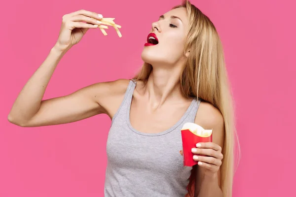 Young beautiful slender girl with a pink background eating a french fries. Concept of un healthy fat junk and healthy food