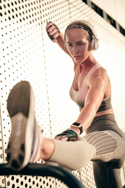 Fitness-Frau mit Kopfhörer beim Stretching und Musikhören vor grauer Wand an sonnigen Tagen, gesunder Lebensstil, Sportkonzept — Stockfoto