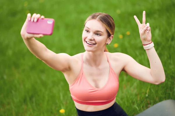 Apto mulher fitness sorridente em sportswear sentado no tapete de ioga na grama verde no parque de verão, tomando selfie no smartphone e mostrando gesto de vitória — Fotografia de Stock