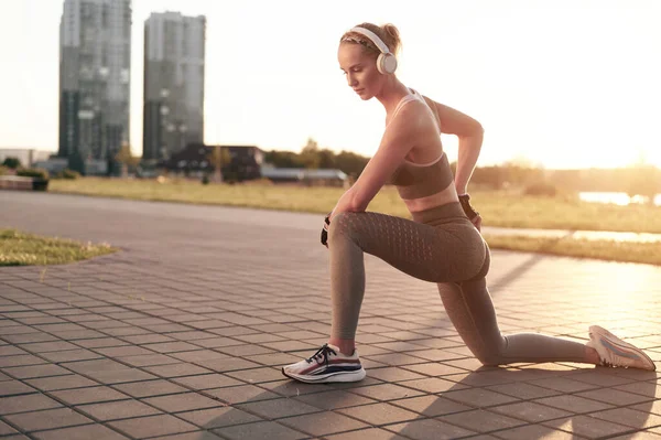 Junge sportliche Frau mit Kopfhörern beim Stretching im Park in der Stadt, kräftige Beine und Bauch, muskulöse Arme, Fitness tragendes Mädchen — Stockfoto