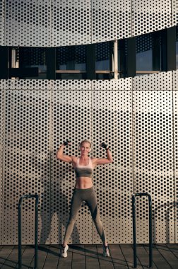 Sıska, kulaklıklı ve spor giyimli genç bir bayan kollarında kaslarını gösteriyor ve müzik dinliyor, güçlü bir kız sokak duvarının yanında poz veriyor.