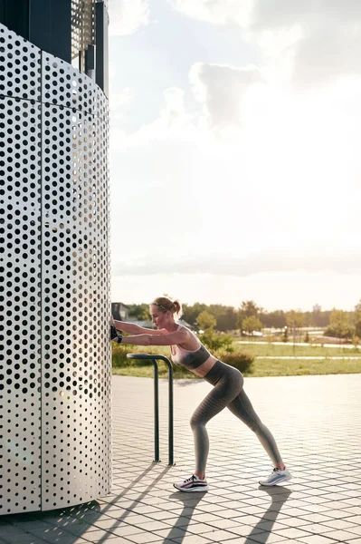 Junge schlanke Fitness-Girl in Sportbekleidung macht körperliche Bewegung vor Gebäude im Freien im Sommerpark, Gegenlicht, Blick auf die Stadt — Stockfoto