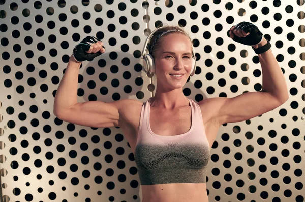 Lächelnde athletische Frau mit Kopfhörern und Sportbekleidung zeigt Muskeln in ihren Armen und hört Musik, starkes Mädchen mit perfektem Körper — Stockfoto