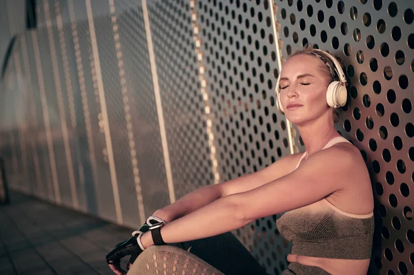 Athletin Läuferin in Sportbekleidung sitzt entspannt und schließt die Augen und hört Musik im Freien vor der Wand — Stockfoto