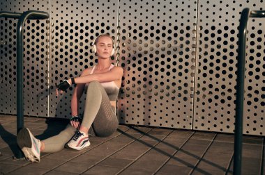 Spor kıyafetli bir kadın duvarın önünde antrenmandan sonra dinleniyor ve kulaklıklarıyla müzik dinliyor.