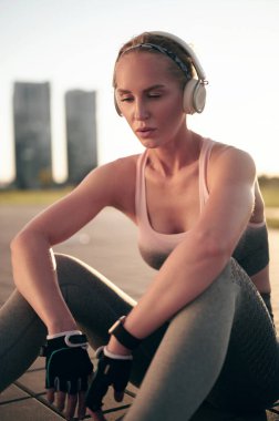 Spor kıyafetleri içinde yorgun ve formda bir kadın fiziksel egzersiz yaparken dinleniyor ve kulaklıkla müzik dinliyor.