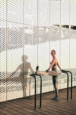 Spor kıyafetleri ve kulaklıklı kadın bina duvarının önünde esneme egzersizleri yapıyor.