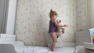 Kollarında bir ayıyla yatakta zıplayan ve eğlenen üç yaşında küçük bir kız.