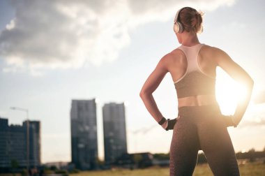 Arka ışık, şehir manzarası, spor giyim bölümündeki güçlü fitness kadını kulaklıkla müzik dinliyor ve dışarıda fiziksel egzersiz yapıyor.