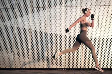 Açık havada egzersiz yapan, koşan ya da zıplayan formda bir kadın. Sağlıklı genç bayan sporcu spor yapıyor ve duvarın önünde kulaklıkla müzik dinliyor.