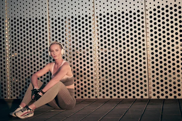 Sportlerin in Fitnessbekleidung sitzt vor Wand, entspannt und hört Musik über Kopfhörer — Stockfoto