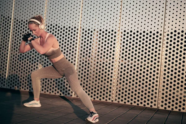 Fit sporty female in fitness wear doing stretching exercise and sit-ups with headphones in front of gray wall — Stock Photo, Image