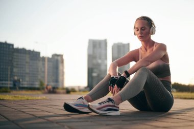 Genç beyaz kadın sporcu antrenman ve fiziksel egzersizden sonra rahatlıyor. Kulaklıkla müzik dinleyen sportif kız. Arka planda gün batımı, şehir manzarası. sağlıklı yaşam tarzı, motivasyon kavramı