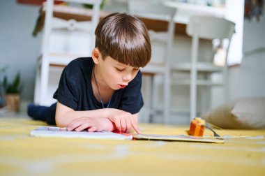 Anaokulu beyaz çocuğu evde yerde uzanıyor, kitap okuyor ve resim seyrediyor. Ev eğitimi kavramı