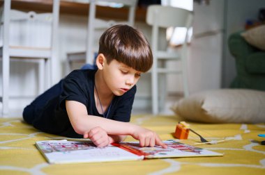 Anaokulu beyaz çocuğu evde yerde uzanıyor, kitap okuyor ve resim seyrediyor. Ev eğitimi kavramı