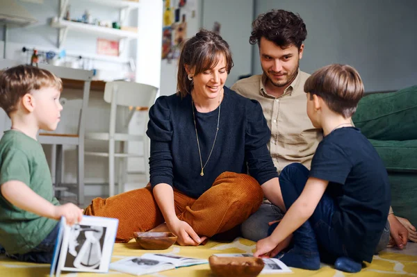 Les jeunes parents caucasiens passent du temps à la maison avec leurs fils et lisent des livres sur le sol. Joyeux jeu de famille avec les enfants d'âge préscolaire. Concept d'enseignement à domicile — Photo