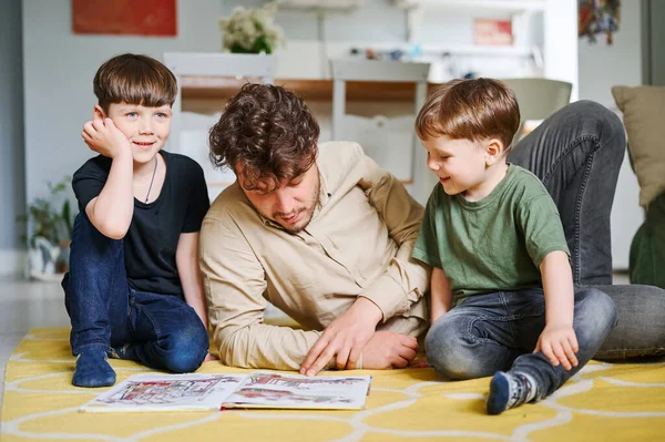 बेटों के साथ पिता किताब पढ़ रहा है, घर पर एक साथ समय बिता रहा है और तल पर पड़ा है — स्टॉक फ़ोटो, इमेज