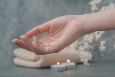 Güzel kadın elleri cilde özen gösterir, krem ve yağ sürerler. Açık renkli bej havlular, spa eşyalar, mumlar ve çiçekler koyu gri arka planda.