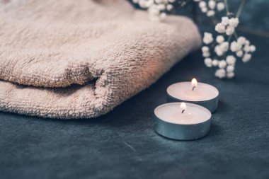 Açık renkli bej havlular, spa eşyalar, mumlar ve çiçekler koyu gri arka planda.