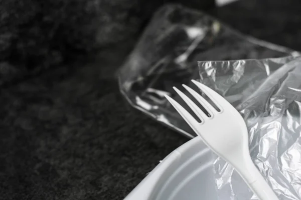 White plastic forks, plate and packages on a gray-black dark background. Plastic waste. Environmental concept. Ban single use plastic. Anti ecology and pollution of the planet