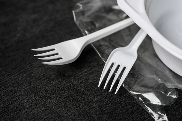 White plastic forks, plate and packages on a gray-black dark background. Plastic waste. Environmental concept. Ban single use plastic. Anti ecology and pollution of the planet