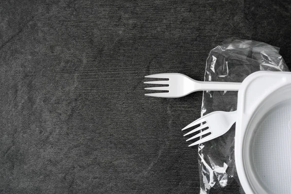 White plastic forks, plate and packages on a gray-black dark background. Plastic waste. Environmental concept. Ban single use plastic. Anti ecology and pollution of the planet