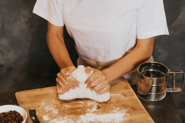 Young figured girl kneads the dough in a white T-shirt with long nails with natural manicure on a gray background. Lush dough with raisins during cooking