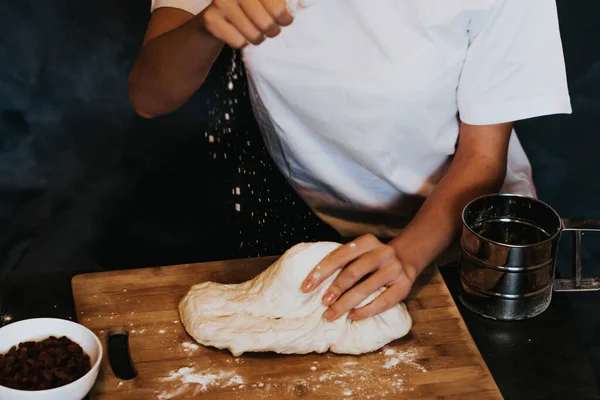 Young figured girl kneads the dough in a white T-shirt with long nails with natural manicure on a gray background. Lush dough with raisins during cooking