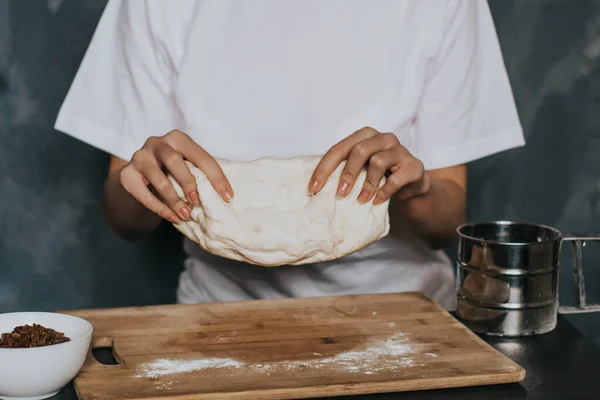 Young figured girl kneads the dough in a white T-shirt with long nails with natural manicure on a gray background. Lush dough with raisins during cooking