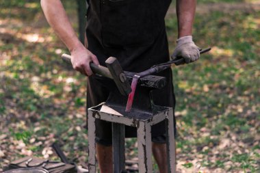 Genç bir demirci doğada bir ürün dövüyor, yanan bir ocak ve küçük taşınabilir bir el örsü. Çekiç işi, metalürji. El yapımı metal. Demirciler Sanatçılık Festivali, Sokak Sergisi.