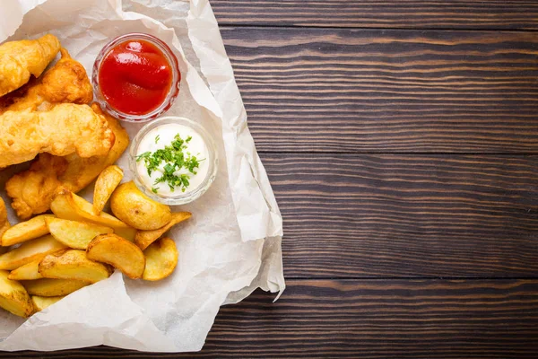 British traditional fast food fish and chips with assorted dips for choice, on paper, rustic wooden background, top view, space for text. Battered fried fish, potato chips, tartare and ketchup sauce